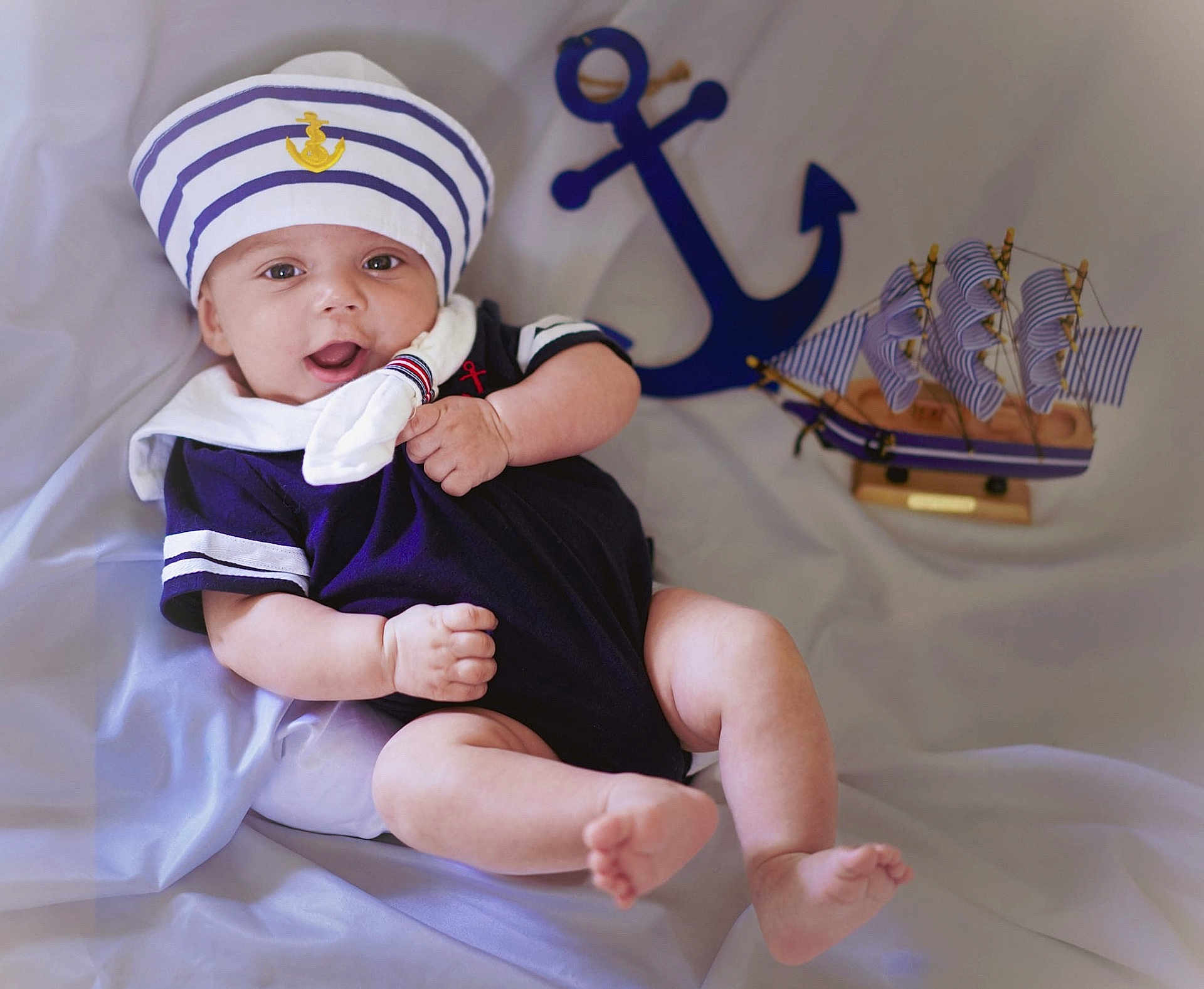 Aaron is registered to the contest to win money with this photo: baby, infant, sailor_hat, navy_outfit, nautical_theme, anchor, toy_ship, smiling, cute, portrait, soft_fabric, indoors, child, happy, skin, arms, legs, headwear, clothing, pose