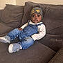 baby, infant, child, denim_overalls, lion_king_beanie, hat, couch, brown_couch, indoor, footwear, socks, long_sleeve_shirt, cute, wide_eyes, seated, person, cozy, soft_furniture, expression, portrait