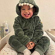 Isaac participe au concours pour gagner de l'argent avec cette photo : child, toddler, smile, hooded_outfit, plush, green, onesie, baby, portrait, indoor, hands, socks, teeth, cute, happy, sitting, nursery, changing_pad, wall, baby_care_products
