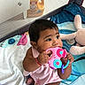 baby, child, infant, sitting, smiling, playpen, toy, toy_phone, pink_toy, plush_bunny, blanket, bedding, indoor, portrait, face, dark_hair, dress, bracelet, nursery, happy