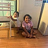 children, toddler, girl, sibling, smiling, portrait, indoor, kitchen, refrigerator, chair, wood_floor, standing, sitting, cross_legged, casual_clothing, pink_shirt, shorts, family, home, playful