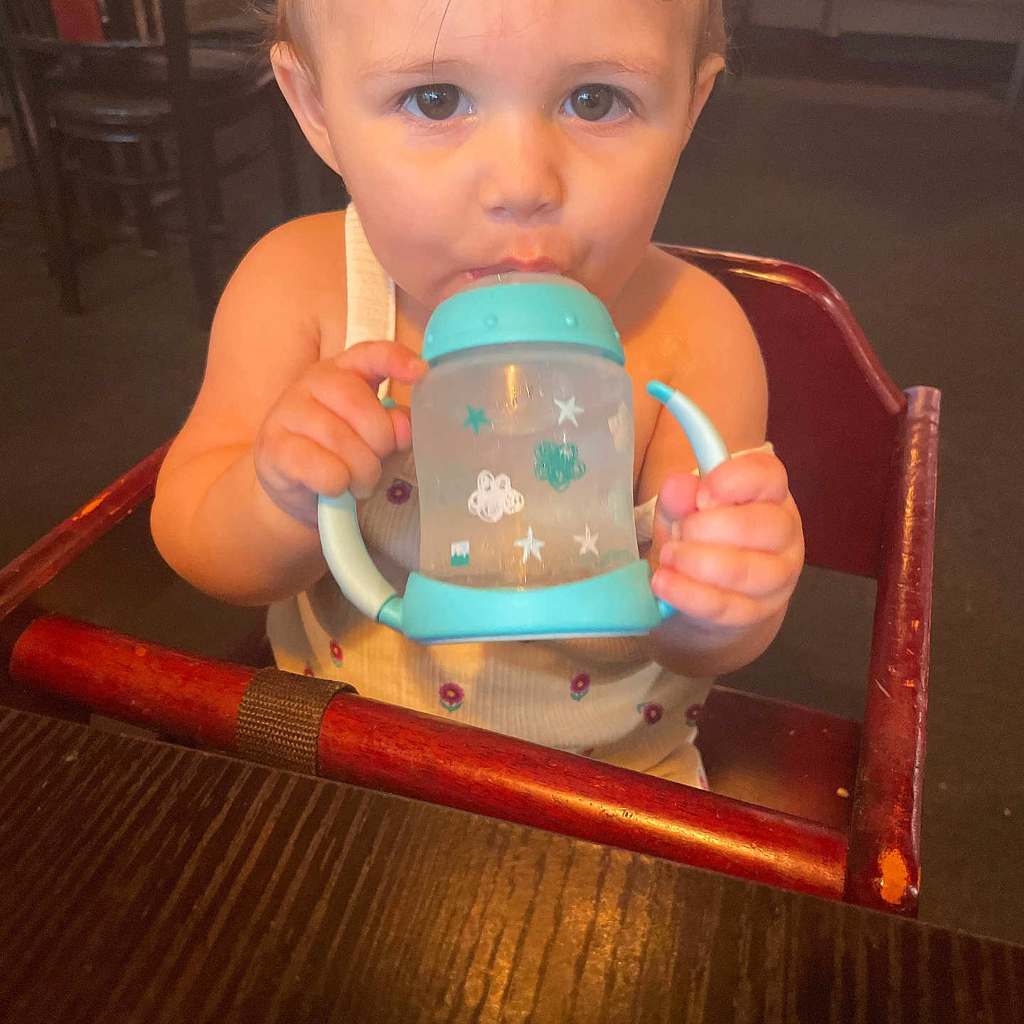Sophia is registered to the contest to win money with this photo: baby, child, close_up, clothing, curious_eyes, cute, drinking, face, furniture, hand, high_chair, indoor, person, seat, sippy_cup, table, toddler, wooden_chair, wooden_table, young_child