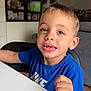 child, boy, smiling, missing_tooth, blue_shirt, sitting, table, chair, indoor, portrait, face, eyes, hair, tooth, happy, home_interior, cabinet, furniture, candid, playful
