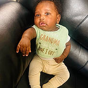 Brayden is registered to the contest to win money with this photo: baby, infant, child, bib, clothing, couch, leather, sitting, reaching, hand, face, expression, cute, indoor, person, sock, pants, soft_lighting, curly_hair, portrait
