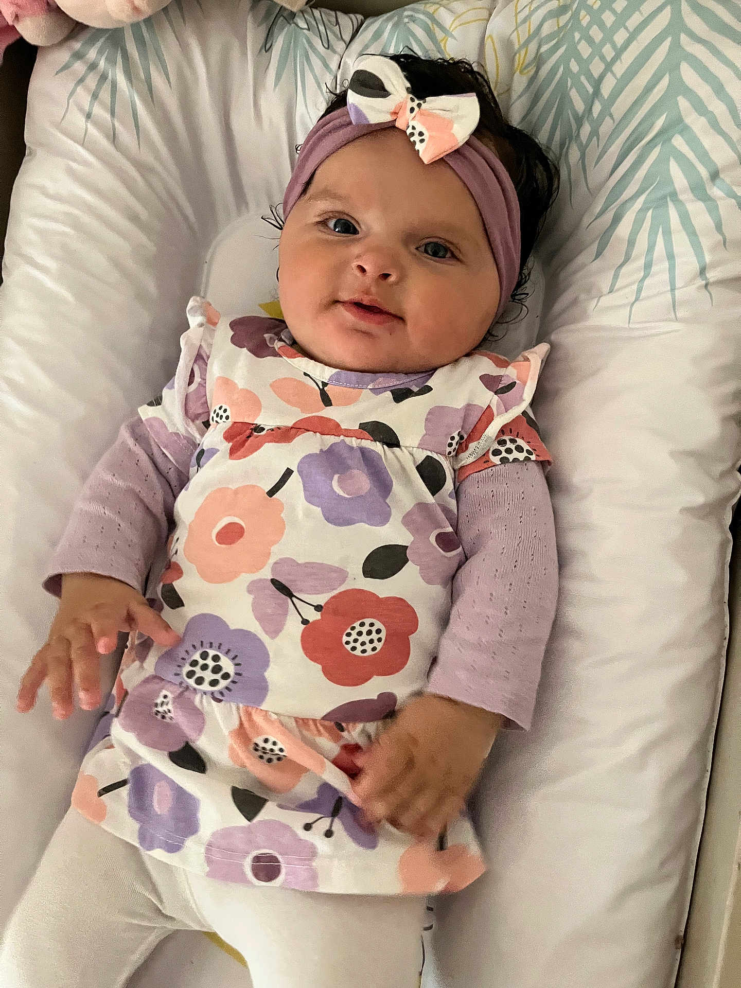 Nalia a rejoint le concours — aidez-le/la à gagner de superbes lots ! baby, child, infant, headband, bow, floral_dress, purple, white_tights, cushion, bedding, lying_down, cute, smiling, portrait, indoor, person, young_child, clothing, hand, face