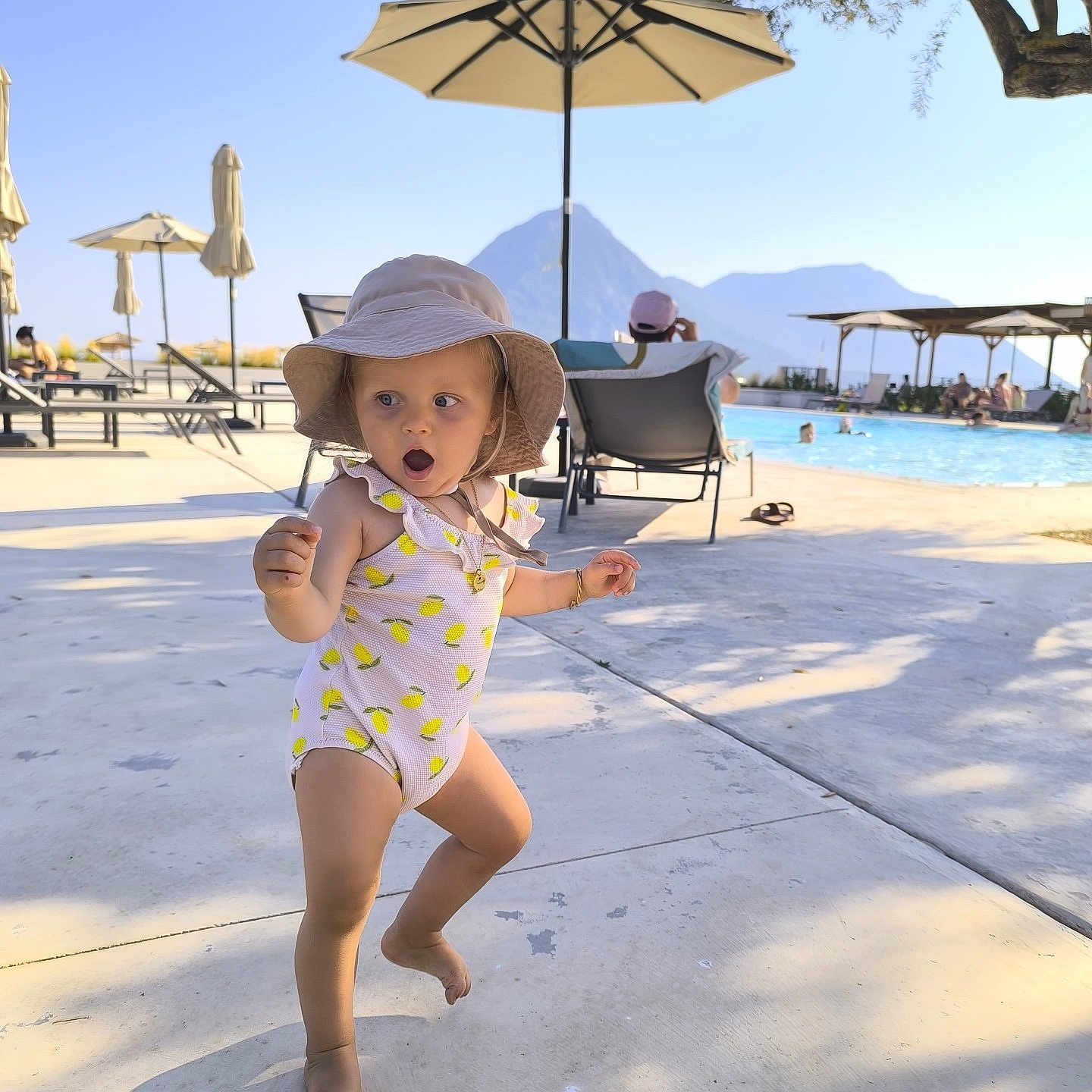 Suzy participe au concours pour gagner de l'argent avec cette photo : barefoot, child, concrete, expression, hat, lounge_chair, mountain, nature, outdoor, person, poolside, recreation, shadow, summer, sunlight, swimsuit, toddler, umbrella, vacation, water
