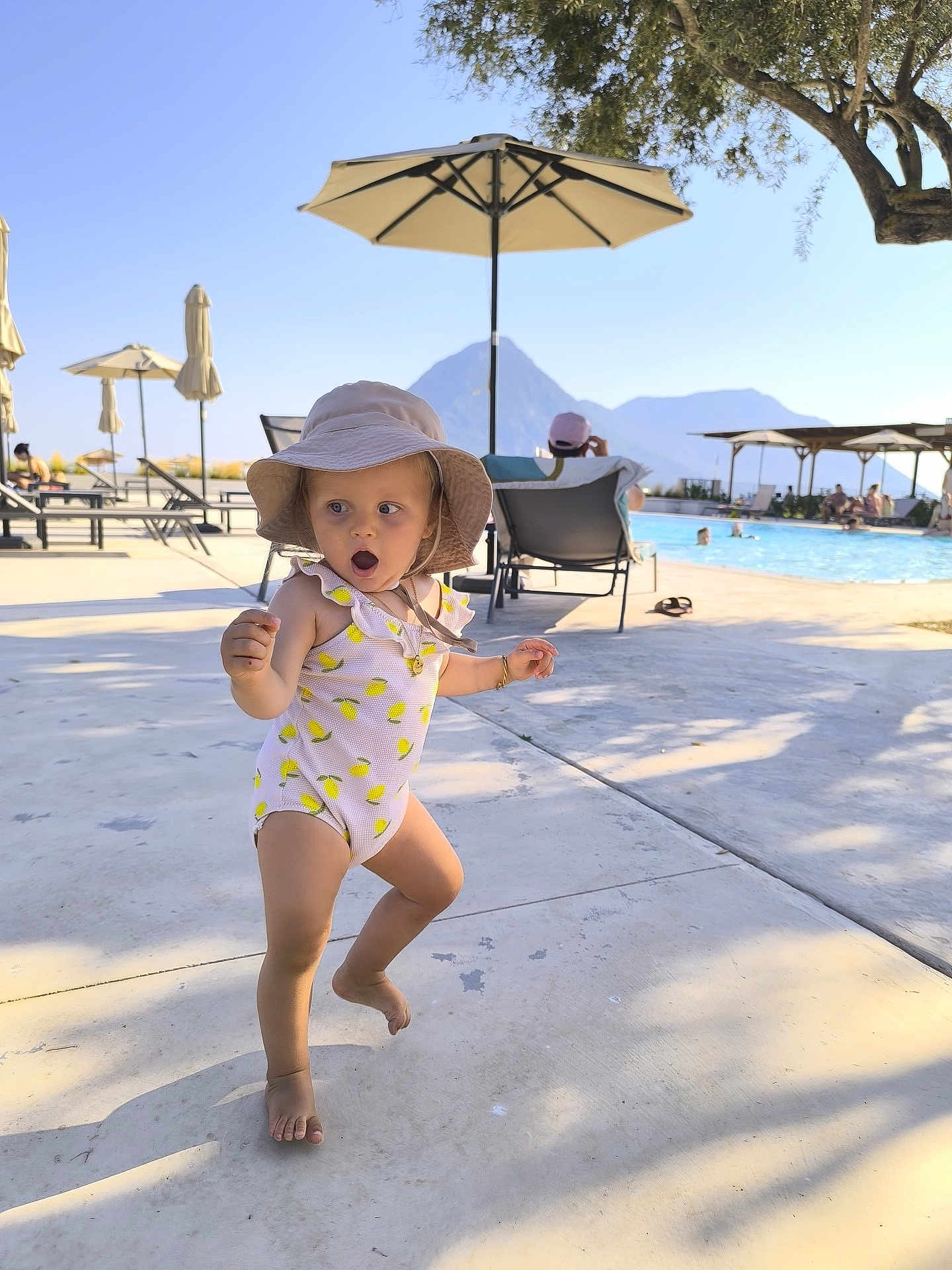 Suzy participe au concours pour gagner de l'argent avec cette photo : toddler, child, swimsuit, hat, poolside, umbrella, mountain, sunlight, shadow, concrete, vacation, outdoor, summer, barefoot, expression, person, water, lounge_chair, recreation, nature
