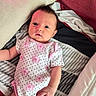 baby, infant, child, portrait, face, eyes, looking_at_camera, lying_down, onesie, floral_pattern, pink_clothing, polka_dots, bed, bedding, pillow, blanket, striped fabric, indoors, close_up, cute