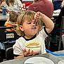 child, toddler, bowl, spoon, eating, restaurant, dining_table, plate, drink, cup, straw, messy_face, person, baby, adult, chair, table, hair, smile, candid