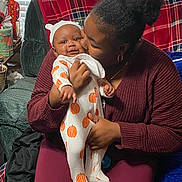 Noah is registered to the contest to win money with this photo: baby, woman, indoor, love, smile, pumpkin_onesie, plaid_blanket, chair, cuddle, family, affection, casual_clothing, headwear, home, window_blinds, hand, face, person, seated, cozy