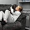 affection, black_dog, cabinet, casual_wear, child, comfort, couch, cozy, dog, floral_wallpaper, furniture, gray_clothing, home, hugging, indoor, living_room, pet, relaxing, sitting, young_person