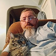 Tucker Brown is registered to the contest to win money with this photo: beard, brown_furniture, casual_clothing, cat, chair, comfort, cuddling, domestic_animal, doorway, facial_hair, glasses, home, indoor, light, man, pet, portrait, relaxing, seated, tabby_cat
