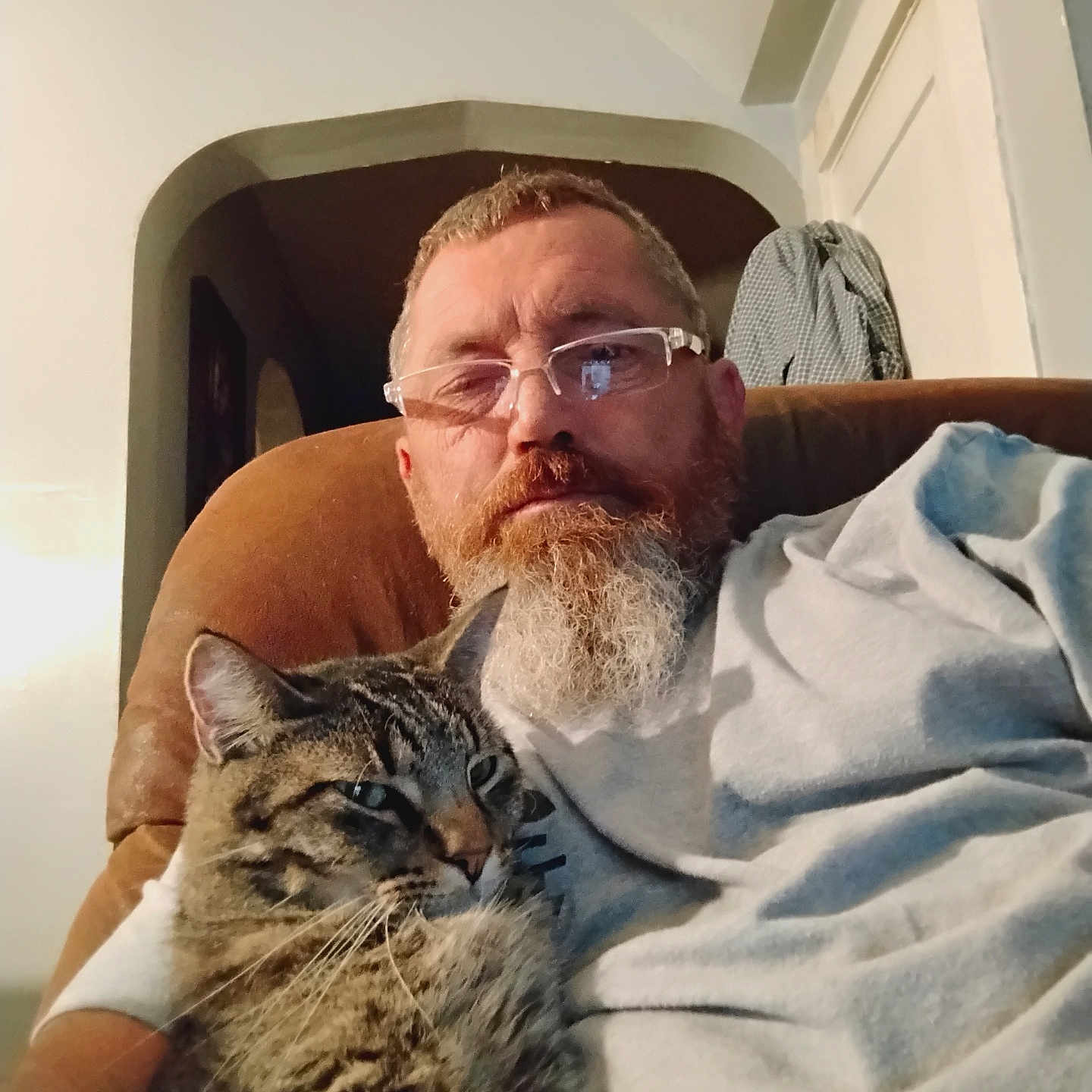 Tucker Brown is registered to the contest to win money with this photo: beard, brown_furniture, casual_clothing, cat, chair, comfort, cuddling, domestic_animal, doorway, facial_hair, glasses, home, indoor, light, man, pet, portrait, relaxing, seated, tabby_cat