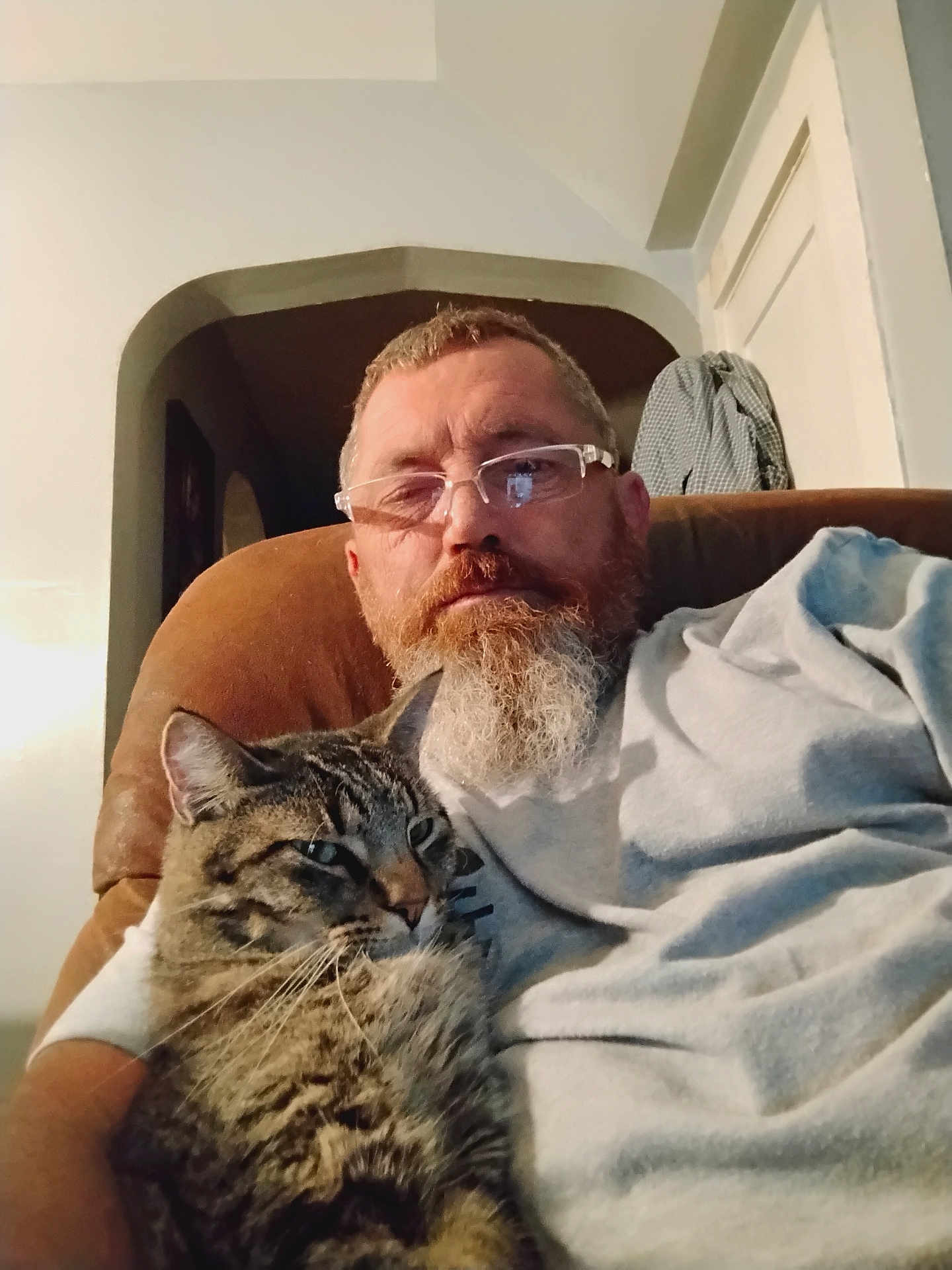 Tucker Brown is registered to the contest to win money with this photo: beard, brown_furniture, casual_clothing, cat, chair, comfort, cuddling, domestic_animal, doorway, facial_hair, glasses, home, indoor, light, man, pet, portrait, relaxing, seated, tabby_cat