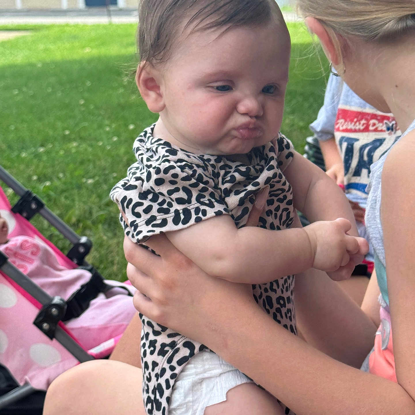 Gracelyn is registered to the contest to win money with this photo: baby, child, pouting, outdoor, grass, stroller, holding, person, diaper, leopard_print, summer, cute, expression, face, portrait, skin, hair, hand, sitting, play