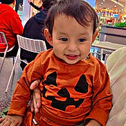 Damian is registered to the contest to win money with this photo: adult, baby, background_people, chair, child, clothing, cute, family, food_court, halloween_outfit, hand, indoor, mall, person, portrait, pumpkin_shirt, signage, smile, table, toddler