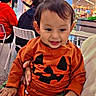 adult, baby, background_people, chair, child, clothing, cute, family, food_court, halloween_outfit, hand, indoor, mall, person, portrait, pumpkin_shirt, signage, smile, table, toddler