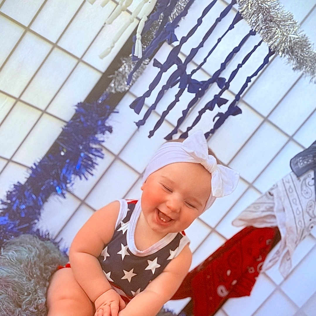 Byrdie is registered to the contest to win money with this photo: baby, bow, child, cute, decorations, festive, fluffy_cushion, happy, headband, holiday, indoor, laughing, onesie, person, portrait, sitting, smiling, star_pattern, streamers, tinsel