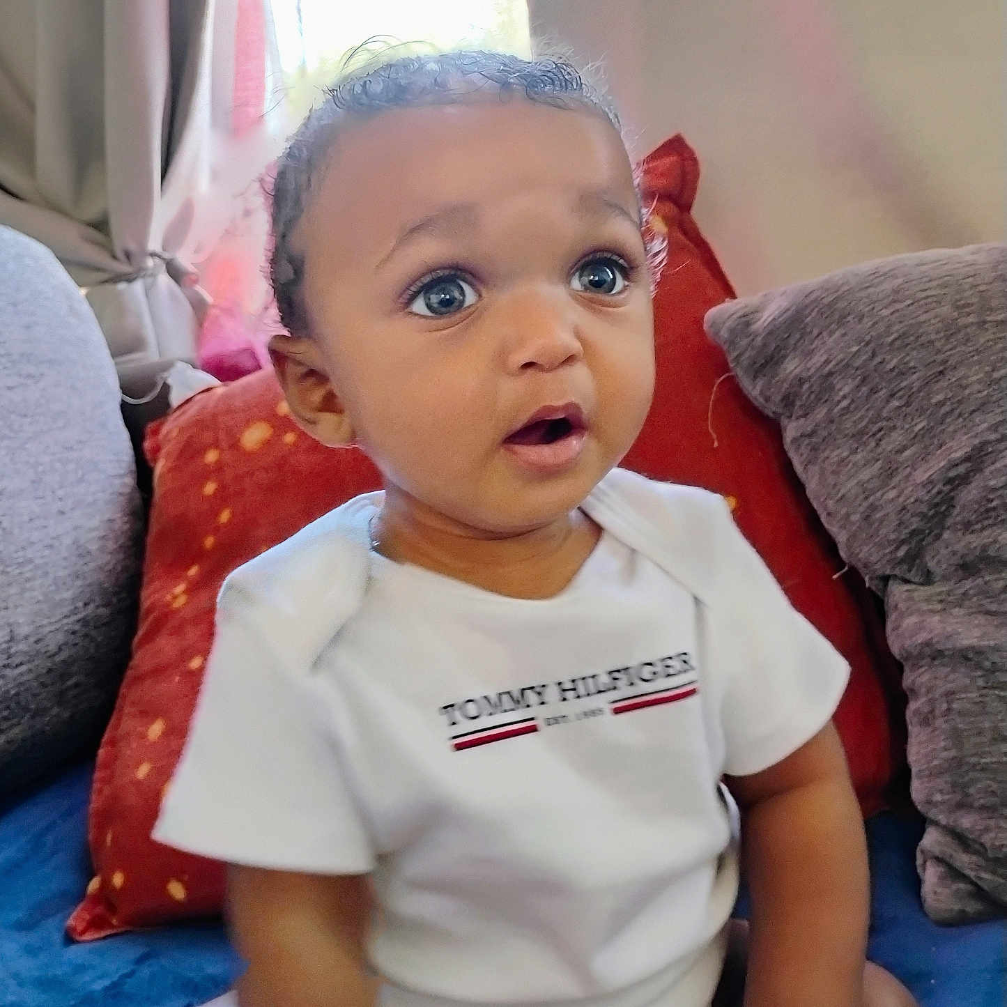 Diego joined the competition — help win amazing prizes! baby, child, infant, person, indoors, couch, pillow, curly_hair, white_clothing, onesie, blue_eyes, expression, seated, home, window, natural_light, red_pillow, gray_pillow, soft_furniture, cute