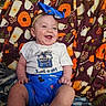 baby, infant, child, smiling, laughing, portrait, indoors, blanket, colorful, bow, headband, white_shirt, blue_shorts, pepsi_logo, sitting, happy, face, person, text_on_clothing, cozy
