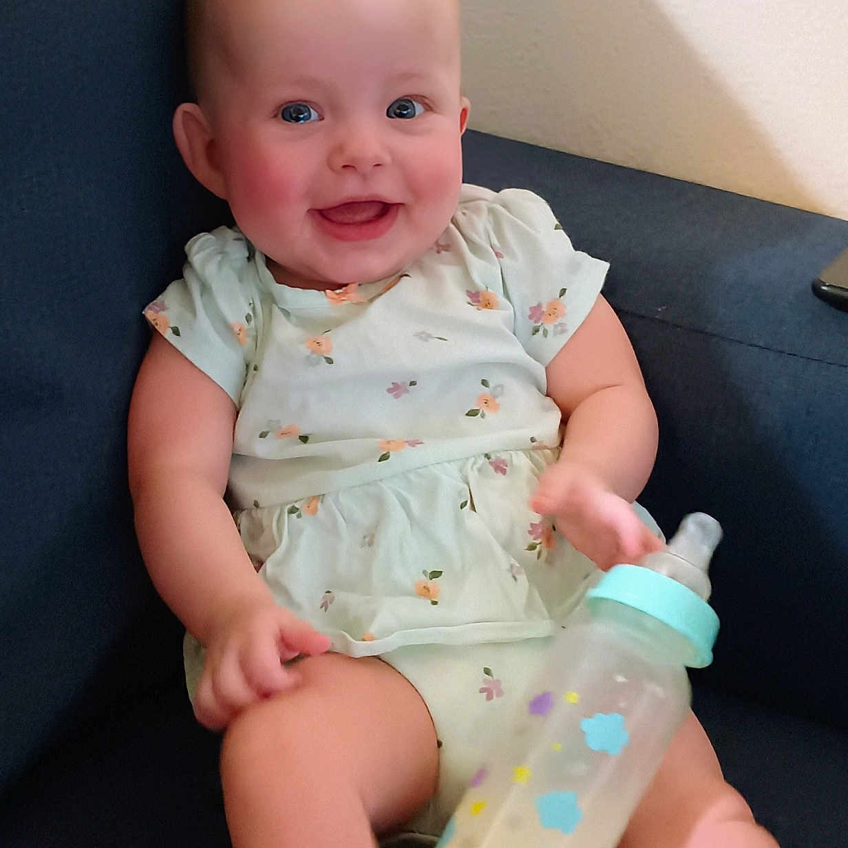 Emelia is registered to the contest to win money with this photo: baby, blue, bottle, chair, child, cute, dress, floral_pattern, happy, indoors, infant, light_skin, milk, person, plush, sitting, smile, soft, toy, young