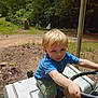 child, toddler, blonde_hair, blue_shirt, green_shorts, brown_boots, steering_wheel, toy_vehicle, outdoor, forest, grass, dirt, summer, play, nature, curious, young_child, vehicle, daylight, casual_clothing