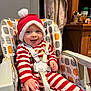 baby, child, smiling, high_chair, santa_hat, holiday, red_stripes, white_stripes, pajamas, indoors, happy, cute, infant, seatbelt, face, person, wooden_furniture, cabinet, toy, colorful