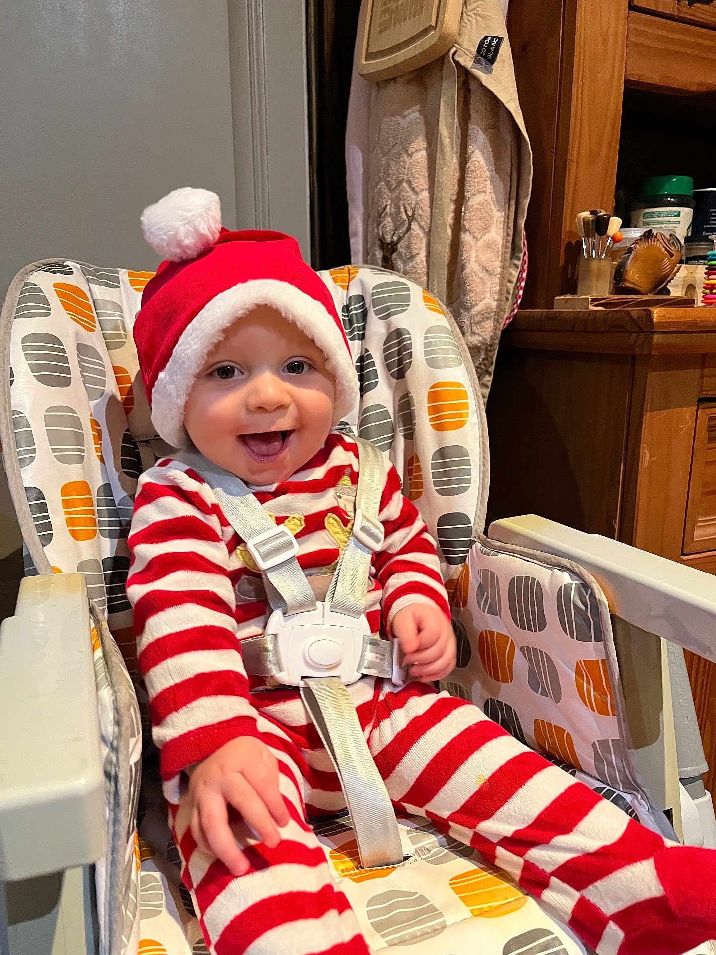 Ayden participe au concours pour gagner de l'argent avec cette photo : baby, child, smiling, high_chair, santa_hat, holiday, red_stripes, white_stripes, pajamas, indoors, happy, cute, infant, seatbelt, face, person, wooden_furniture, cabinet, toy, colorful