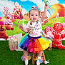 child, toddler, rainbow_tutu, colorful_clothing, lollipop, smiling, happy, indoor, grass_carpet, candyland_theme, background, gingerbread_man, cupcake, toy, festive, party, cute, socks, ribbon, wooden_stool