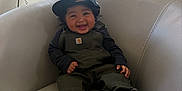 Jax is registered to the contest to win money with this photo: toddler, child, smiling, sitting, overalls, cap, sneakers, couch, indoor, window, happy, person, fashion, footwear, portrait, curved_furniture, natural_light, casual_clothing, black_hair, cute