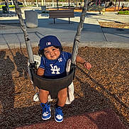 Jax joined the competition — help win amazing prizes! toddler, child, swing, playground, cap, blue_clothing, sneakers, smile, outdoor, wood_chips, shade, bench, trees, sunlight, play_area, fence, concrete, happy, baby_swing, park