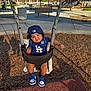 toddler, child, swing, playground, cap, blue_clothing, sneakers, smile, outdoor, wood_chips, shade, bench, trees, sunlight, play_area, fence, concrete, happy, baby_swing, park