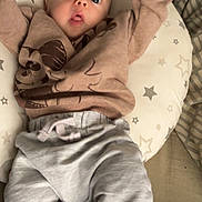 Archie is registered to the contest to win money with this photo: baby, infant, cushion, star_pattern, sweatshirt, pants, raised_arms, wide_eyes, surprised_expression, indoor, fabric, soft_toys, bed, sleeping_area, child, person, cute, cozy, casual_clothing, relaxed