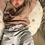baby, infant, cushion, star_pattern, sweatshirt, pants, raised_arms, wide_eyes, surprised_expression, indoor, fabric, soft_toys, bed, sleeping_area, child, person, cute, cozy, casual_clothing, relaxed