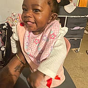 Kaylani is registered to the contest to win money with this photo: baby, child, smiling, indoor, bib, pink, hair_bows, happy, person, infant, clothing, holding, tattoo, arm, floor, furniture, toy, footwear, cute, young