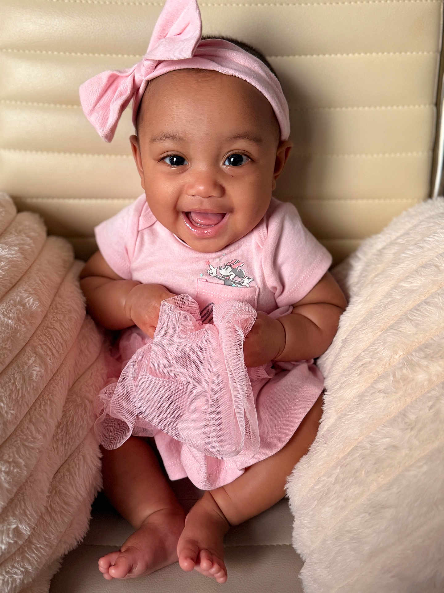 Leora is registered to the contest to win money with this photo: baby, child, pink_clothing, headband, smiling, happy, cushion, fabric, sitting, cute, indoor, infant, skin, face, hands, feet, soft_texture, comfort, portrait, closeup