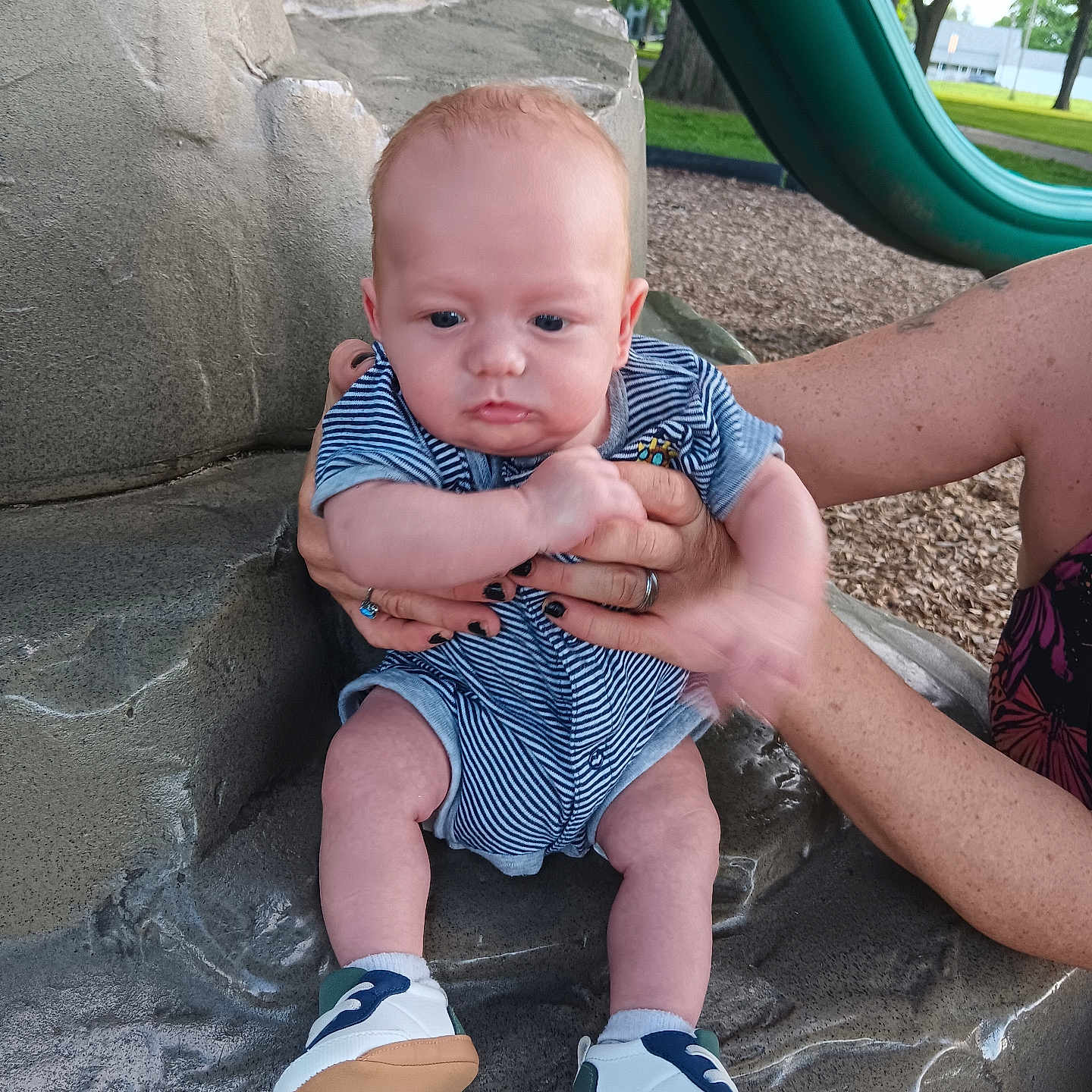 Allan is registered to the contest to win money with this photo: baby, infant, child, striped_clothing, blue_onesie, shoes, hands, adult, playground, slide, stone_surface, outdoor, park, grass, trees, daylight, skin, person, sitting, cute