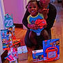 child, adult, basketball, toys, gifts, floor, indoor, wood_floor, smiling, person, black_hair, colorful, puzzle, snack, packaging, celebration, home, walls, doorway, happy