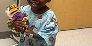 Chase is registered to the contest to win money with this photo: child, smiling, hospital_gown, stuffed_toy, tiger, medical_equipment, hospital_room, door, floor, phone, indoor, person, healthcare, young_child, toy, happy, medical_device, childrens_clothing, portrait, standing