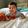 baby, smiling, bed, blanket, infant, child, happy, indoors, onesie, fabric, face, person, cute, crib, colorful_cloth, home, portrait, young_child, lying_down, soft_lighting