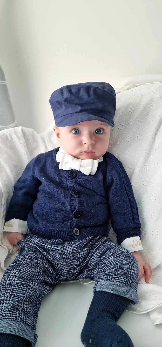 Gabriel is registered to the contest to win money with this photo: baby, child, blue_eyes, navy_blue_hat, navy_blue_cardigan, white_bow_tie, checkered_pants, socks, sitting, white_blanket, curious_expression, indoor, portrait, clothing, infant, cute, fashion, young_child, face, person
