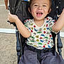 toddler, stroller, child, smiling, happy, shorts, shirt, insect_pattern, outdoor, concrete, person, walking, legs, black_stroller, seatbelt, joyful, arm_raised, casual, daylight, cute