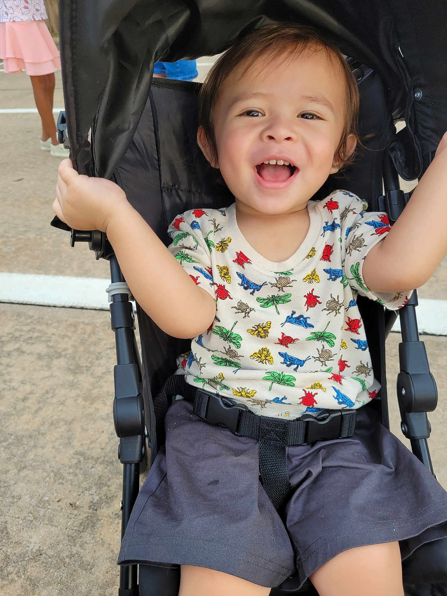 Leo joined the competition — help win amazing prizes! toddler, stroller, child, smiling, happy, shorts, shirt, insect_pattern, outdoor, concrete, person, walking, legs, black_stroller, seatbelt, joyful, arm_raised, casual, daylight, cute