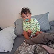 Jacquelyn joined the competition — help win amazing prizes! toddler, child, bed, pillow, blanket, pacifier, curly_hair, diaper, yellow_shirt, patterned_clothing, indoor, casual, sitting, looking_away, cozy, relaxed, soft_lighting, home, portrait, young_child