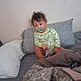 toddler, child, bed, pillow, blanket, pacifier, curly_hair, diaper, yellow_shirt, patterned_clothing, indoor, casual, sitting, looking_away, cozy, relaxed, soft_lighting, home, portrait, young_child