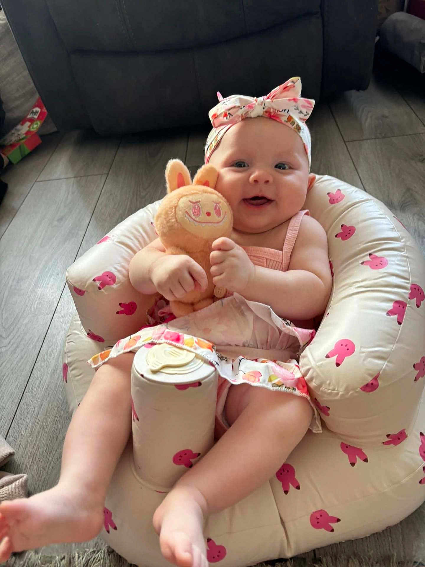 Darcie-Rae joined the competition — help win amazing prizes! baby, infant, child, plush_toy, pink, headband, smiling, sitting, indoor, flooring, inflatable_chair, bunny_pattern, cute, happy, toy, dress, footwear_free, living_room, soft_lighting, playful