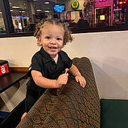 Legend is registered to the contest to win money with this photo: toddler, child, smiling, curly_hair, lollipop, diner, booth, patterned_seat, indoor, neon_sign, reflection, casual_clothing, black_shirt, happy, young_child, person, table, restaurant, candid, portrait