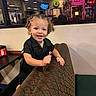 toddler, child, smiling, curly_hair, lollipop, diner, booth, patterned_seat, indoor, neon_sign, reflection, casual_clothing, black_shirt, happy, young_child, person, table, restaurant, candid, portrait