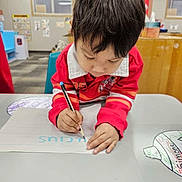 Lucius is registered to the contest to win money with this photo: child, writing, paper, table, red_sweater, classroom, indoor, drawing, marker, focus, young_child, education, learning, hand, person, desk, colored_paper, cute, school, activity