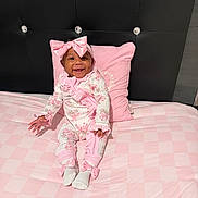Journi is registered to the contest to win money with this photo: baby, infant, smiling, pink, bow, pajamas, bed, pillow, headboard, checkered, floral, clothing, cute, happy, sitting, indoors, portrait, child, person, soft